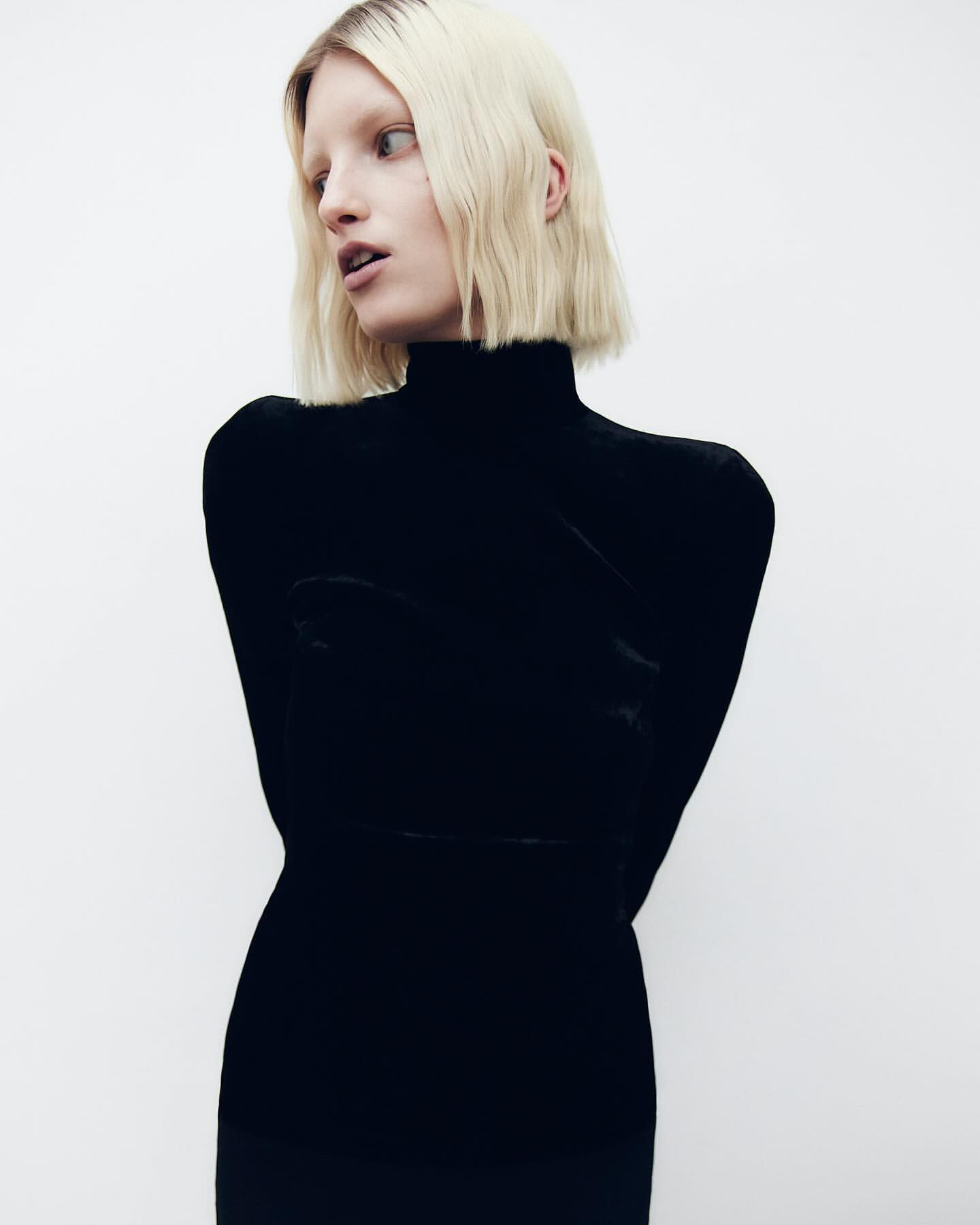 A fashion portrait of a blonde woman wearing a black turtleneck dress against a white backdrop in a studio setting.