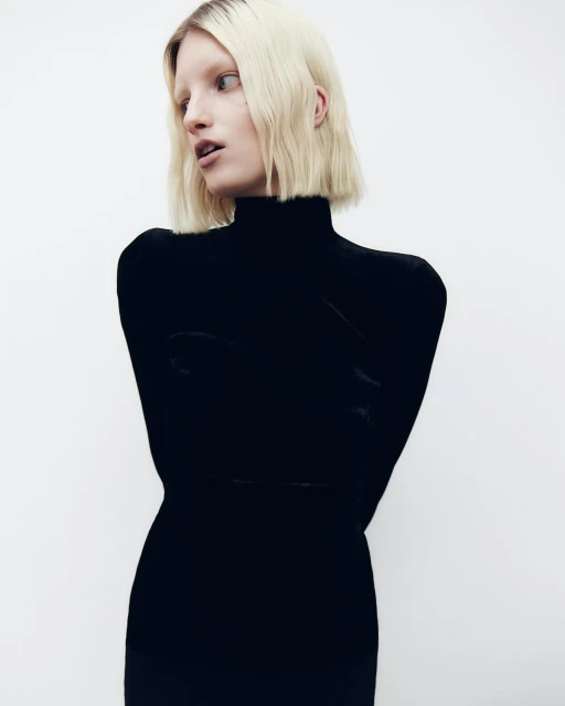 A fashion portrait of a blonde woman wearing a black turtleneck dress against a white backdrop in a studio setting.