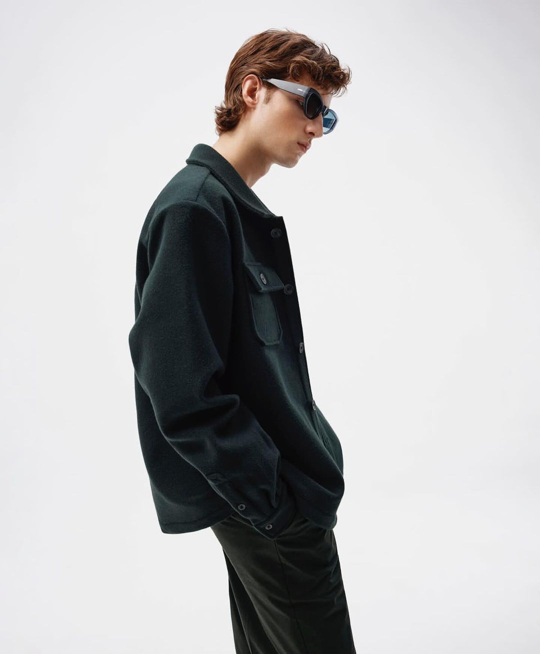 Male fashion model wearing dark green shirt jacket and dark pants against white backdrop in studio setting with sunglasses.