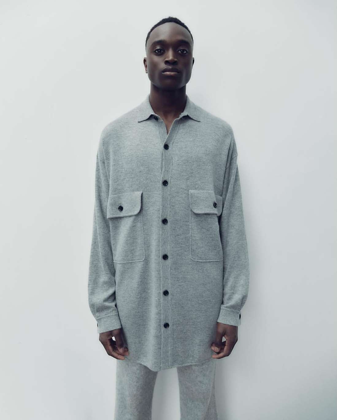 Fashion shot of a Black male model in a gray button-down shirt and matching pants against a neutral backdrop, showcasing minimalist style.