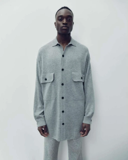 Fashion shot of a Black male model in a gray button-down shirt and matching pants against a neutral backdrop, showcasing minimalist style.
