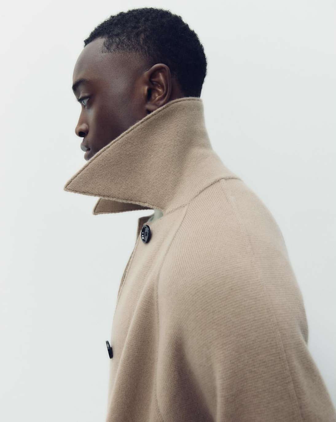 Profile shot of a Black male in a tan coat with a high collar, against a white background, emphasizing fashion and minimalist aesthetic.