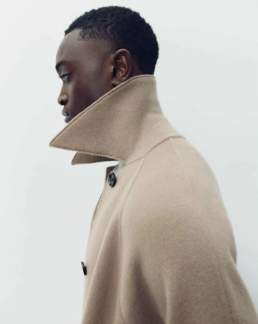 Profile shot of a Black male in a tan coat with a high collar, against a white background, emphasizing fashion and minimalist aesthetic.