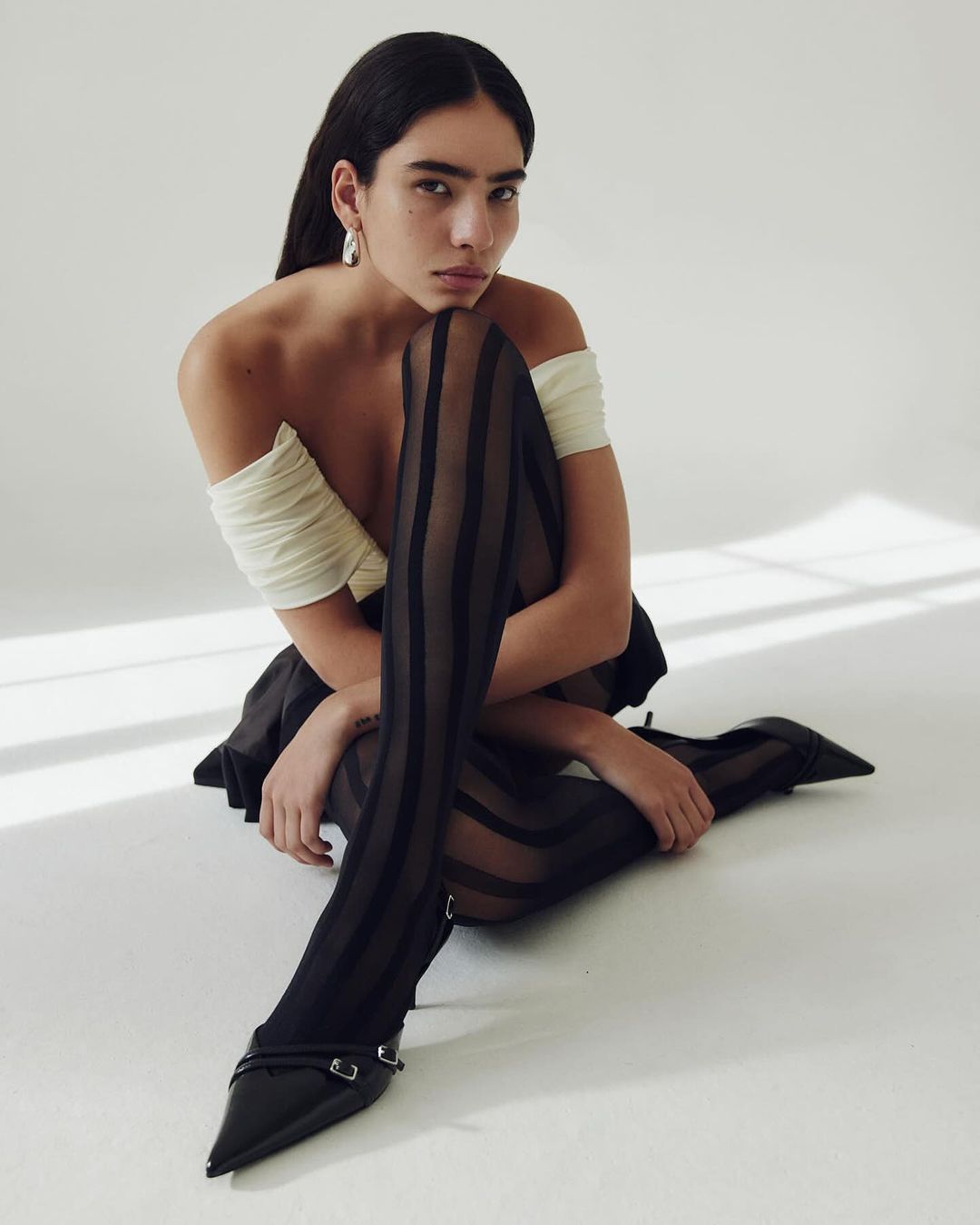 A young adult woman poses sitting on the floor with legs crossed, wearing a cream top, black skirt, and black striped tights with heels in a studio.