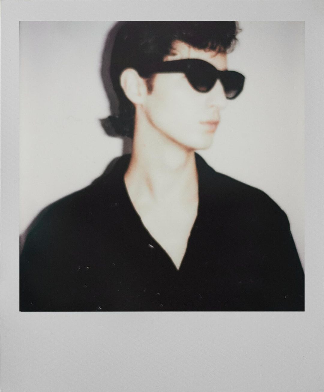 Portrait of a person wearing black sunglasses and a black button-down shirt, set against a neutral background, captured with soft lighting.
