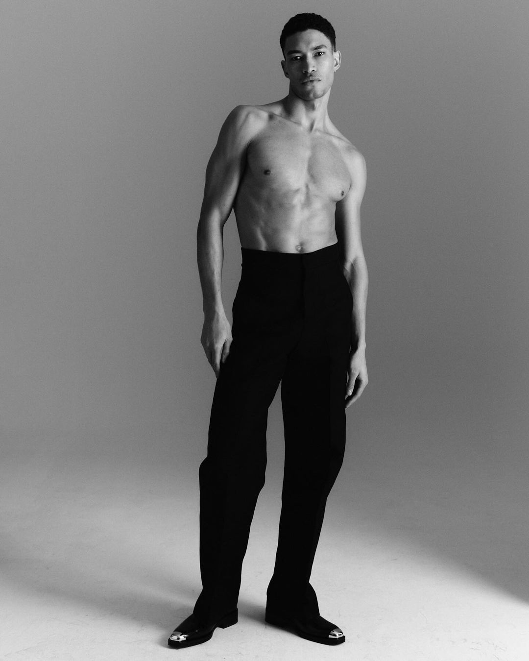 Striking black and white portrait of a muscular adult male in black pants posing in a studio with a neutral grey background.