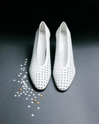 A pair of white perforated high heel pumps placed on a dark grey surface next to scattered confetti and small orange pills.