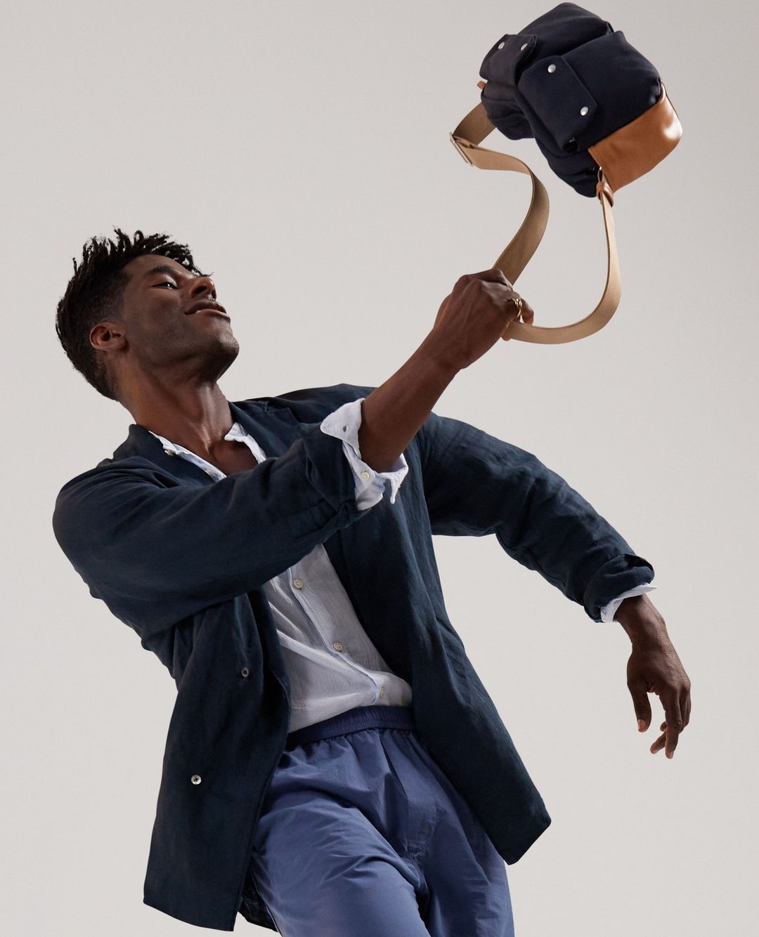 Man in blue linen suit throws blue canvas and leather satchel in a light studio. Fashion editorial featuring modern menswear.