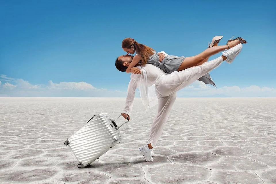 A man carrying a woman in his arms with a suitcase on a cracked plain under a bright sky, embodying travel and lifestyle.