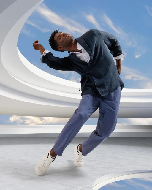 Man in contemporary dance pose in casualwear, against architectural background with blue sky and clouds, captured in a full shot.