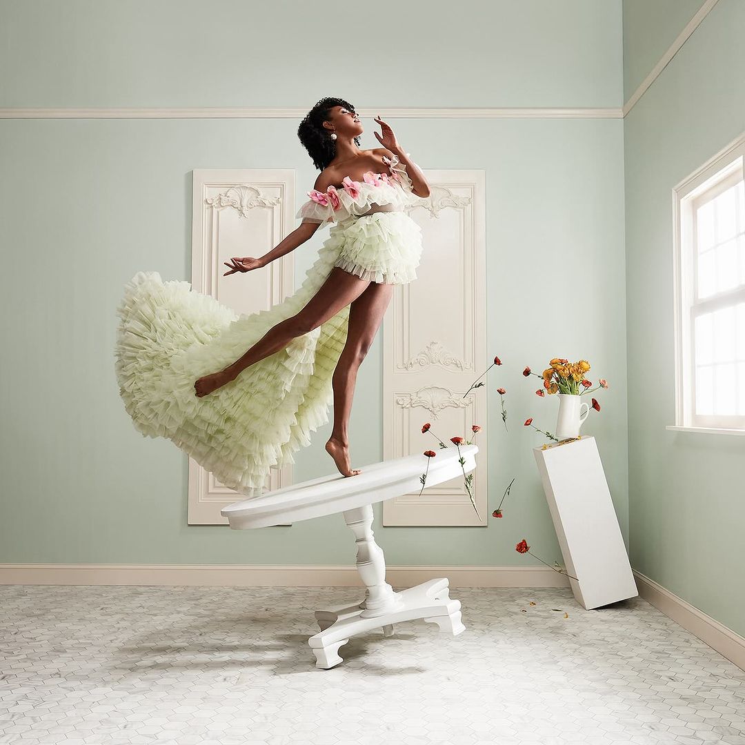 An African American woman in an elaborate, light green dress dances on a white table in a room with light blue walls and white trim.