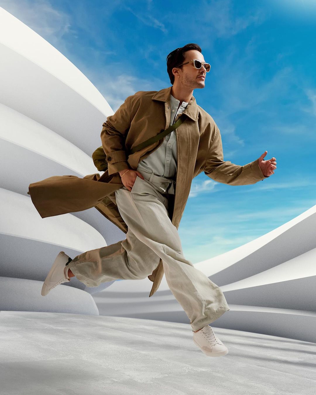 A man in beige clothing and sunglasses runs in front of a white structure against a blue sky, emphasizing fashion and movement.