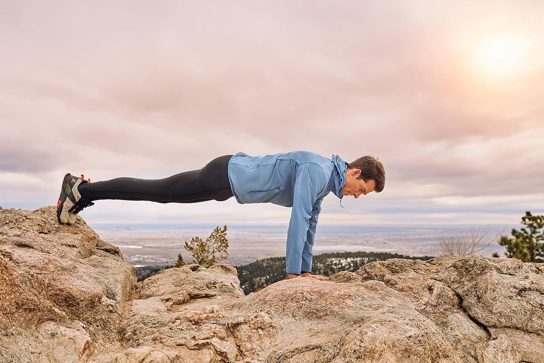 Athletic man in blue jacket performing a plank exercise on a rocky mountain peak with a scenic landscape and sky in the background.