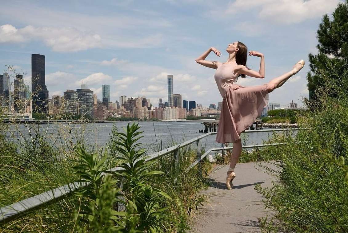 Ballet dancer poses en pointe outdoors with a river and city skyline backdrop in a graceful dance, captured in natural light.