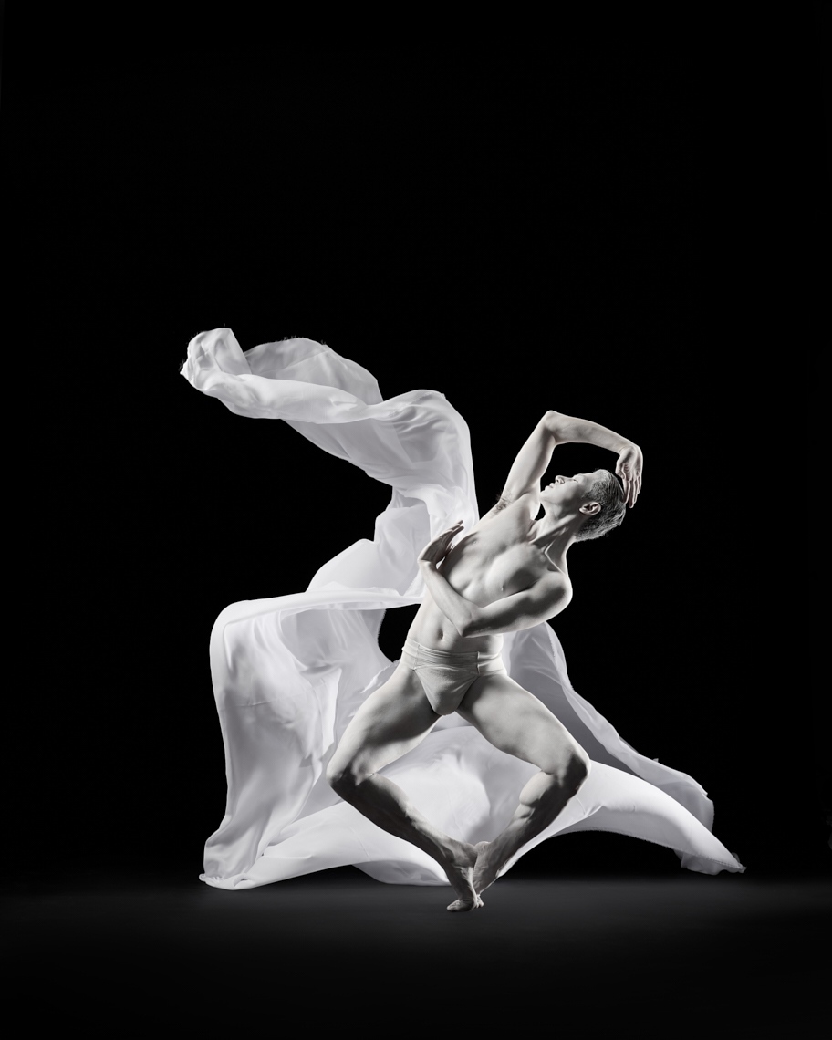 Monochromatic artistic studio shot of a muscular adult male dancer in dynamic pose with white flowing fabric on a dark background.