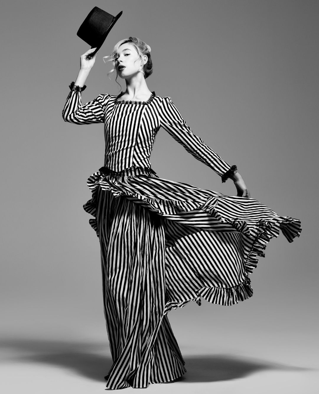 Woman in a striped dress holds a black top hat, posing in a studio. Black and white fashion portrait with neutral grey background.