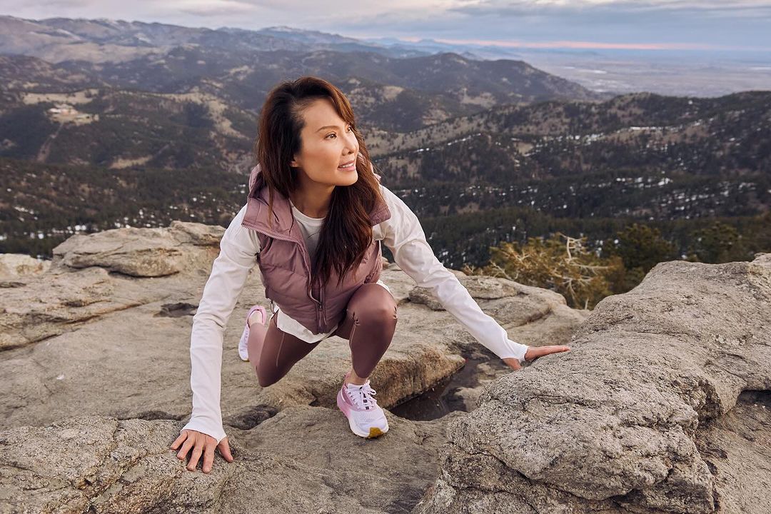 Adult woman on a mountain peak in athletic wear pauses while hiking to enjoy the view on a sunny day with mountains in the background.