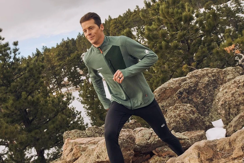 A man in activewear runs on a rocky, snow-dusted trail in a mountain landscape, wearing a green jacket and black pants on a sunny day.