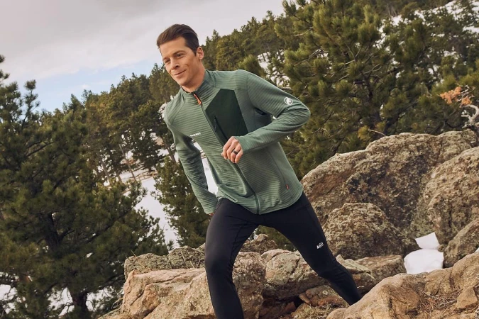 A man in activewear runs on a rocky, snow-dusted trail in a mountain landscape, wearing a green jacket and black pants on a sunny day.