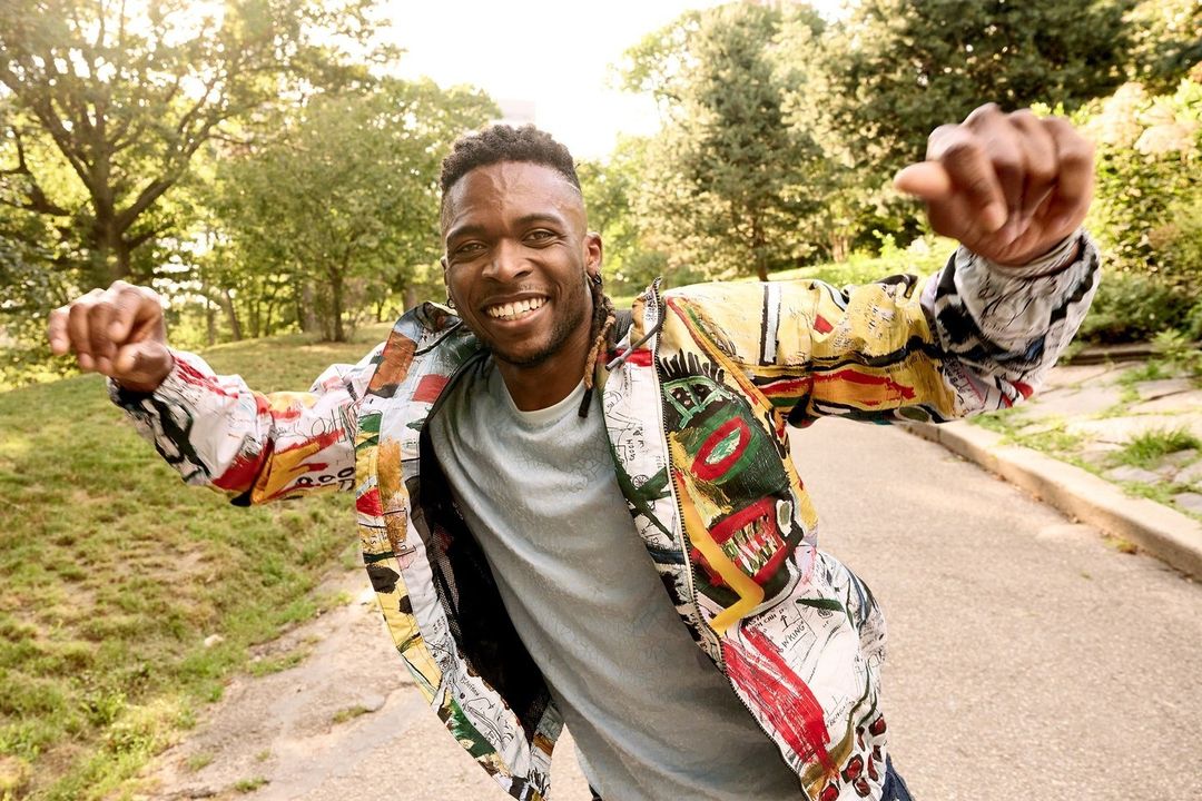 A smiling adult male in patterned jacket stands outdoors on pathway surrounded by green trees and grass, bathed in soft natural light.