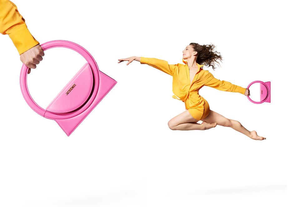 A fashion shot showcases a pink Jacquemus handbag being passed between two people in yellow outfits against a white background.