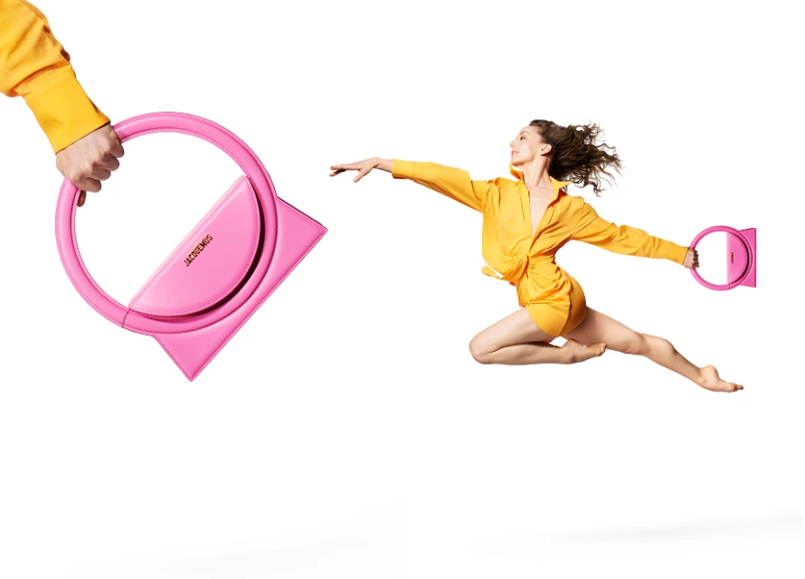 A fashion shot showcases a pink Jacquemus handbag being passed between two people in yellow outfits against a white background.