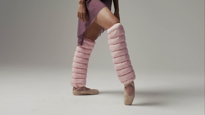 Ballet dancer stands en pointe with pink padded leg warmers and pointe shoes in a modern studio setting with soft, neutral lighting.