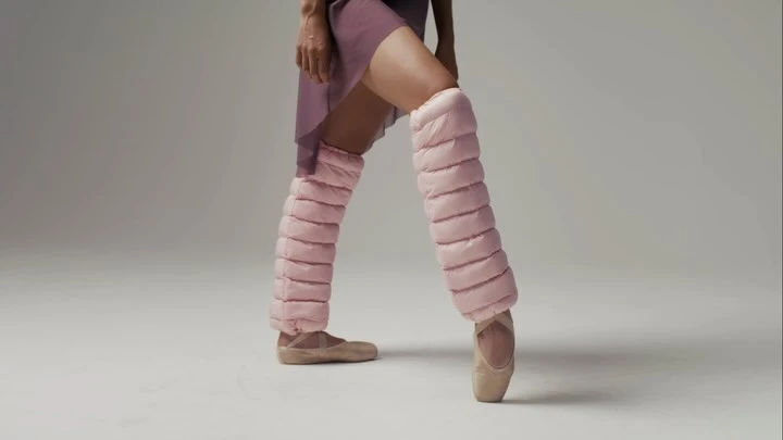 Ballet dancer stands en pointe with pink padded leg warmers and pointe shoes in a modern studio setting with soft, neutral lighting.