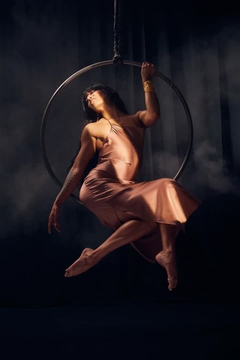 A woman in a peach-colored dress gracefully performs aerial acrobatics on a suspended metal ring, set against a dramatic, dark background.