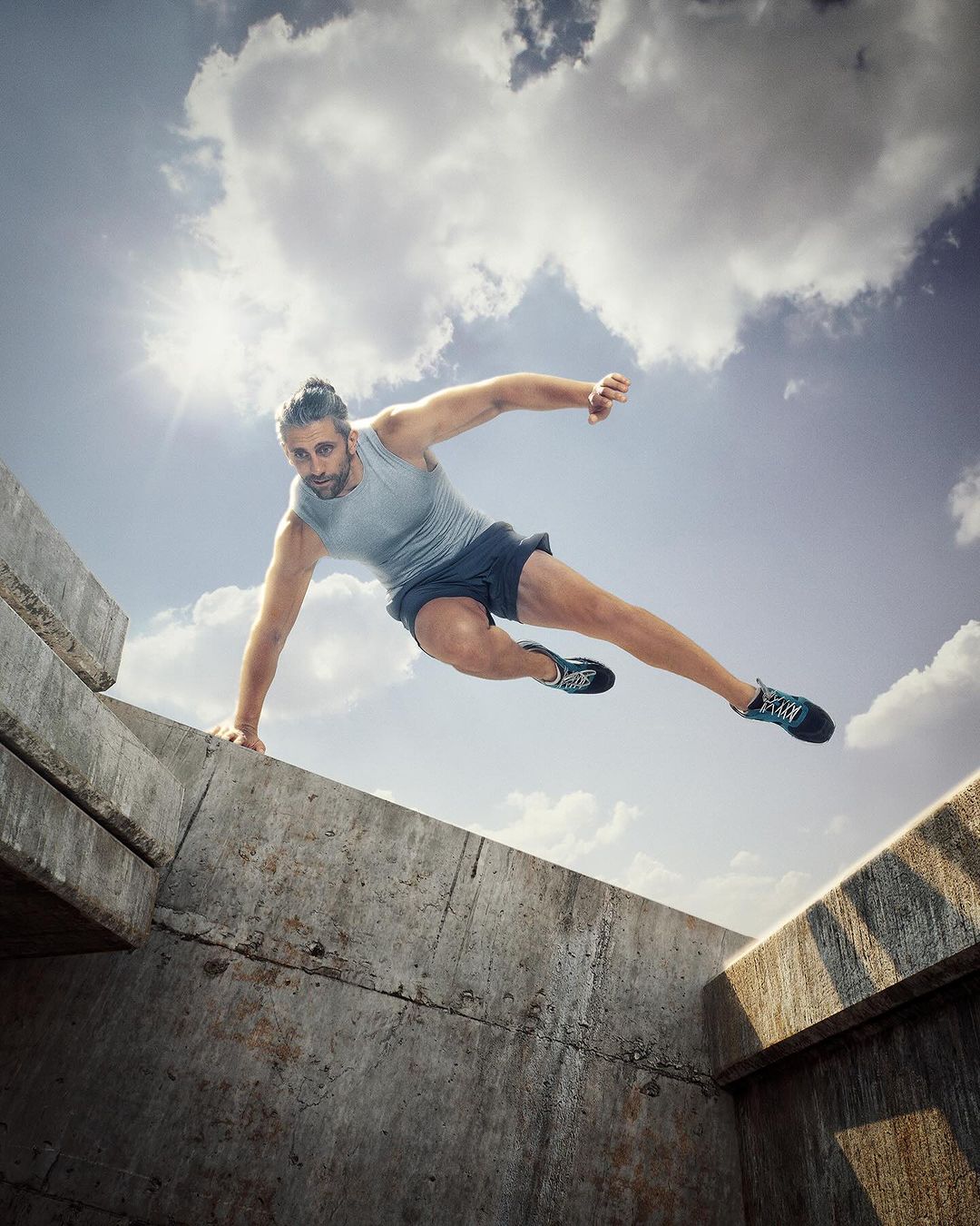 Athletic man mid-air, leaping over concrete structures against a cloudy sky, captured with a dynamic, low-angle perspective, showcasing fitness and agility.