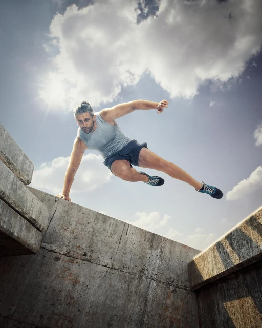 Athletic man mid-air, leaping over concrete structures against a cloudy sky, captured with a dynamic, low-angle perspective, showcasing fitness and agility.