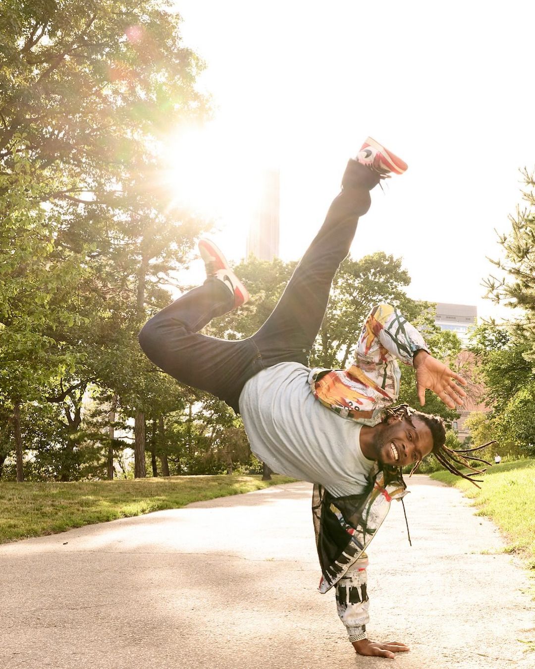 Male dancer breakdancing on one hand on a park path, wearing athletic clothing in an urban park setting with sunlight.