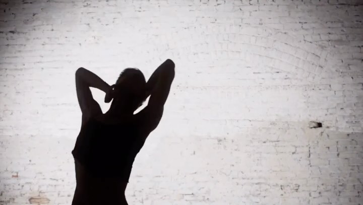 Silhouette of a slender adult female in a sports bra and yoga pants against a textured white brick wall, hands behind her head.