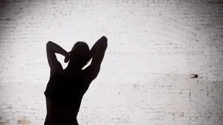 Silhouette of a slender adult female in a sports bra and yoga pants against a textured white brick wall, hands behind her head.