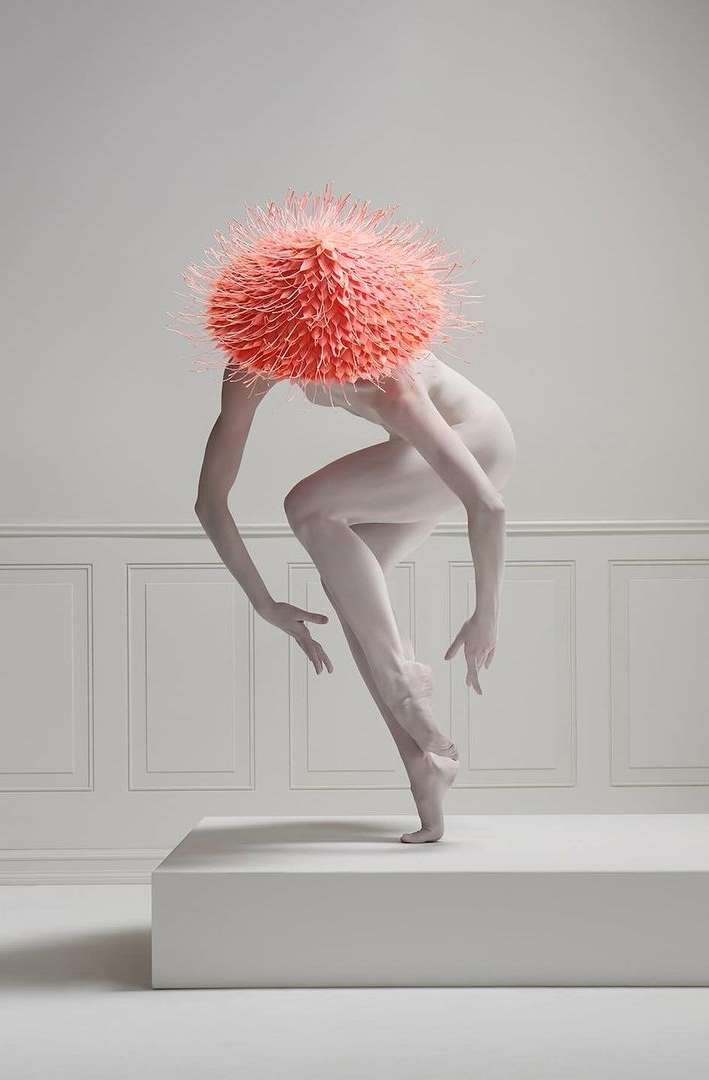 Artistic studio shot of a dancer with white body paint and a large orange headpiece, striking a ballet pose on a white pedestal.