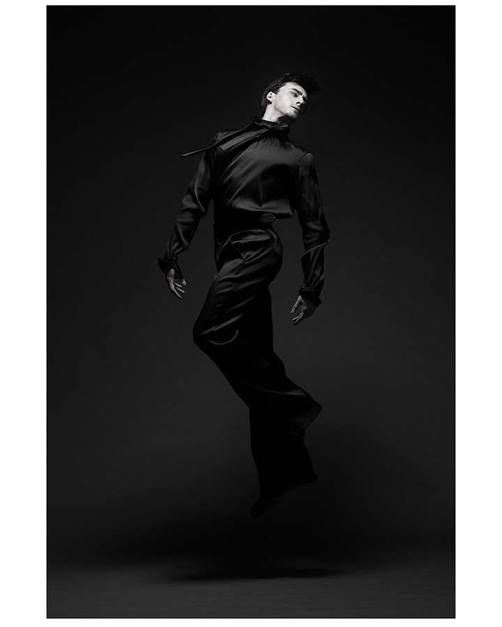 Monochrome studio shot of a young adult male model in mid-air, wearing a black satin long-sleeve shirt and wide-leg pants, leaping.
