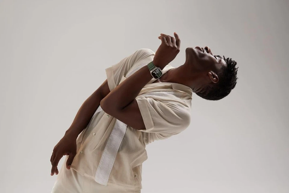 Dark-skinned man in light shirt with smartwatch bends backward in a bright studio with soft lighting, showcasing fashion and technology.