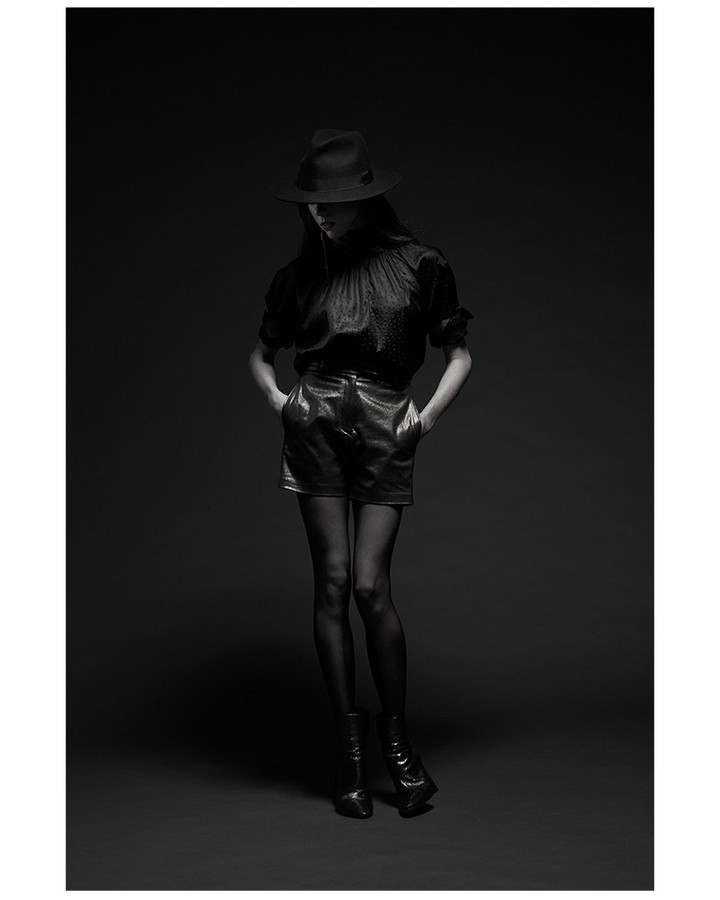 Monochrome studio shot of a female model in a hat and dark, shiny blouse and shorts, wearing tights and heeled boots against dark backdrop.