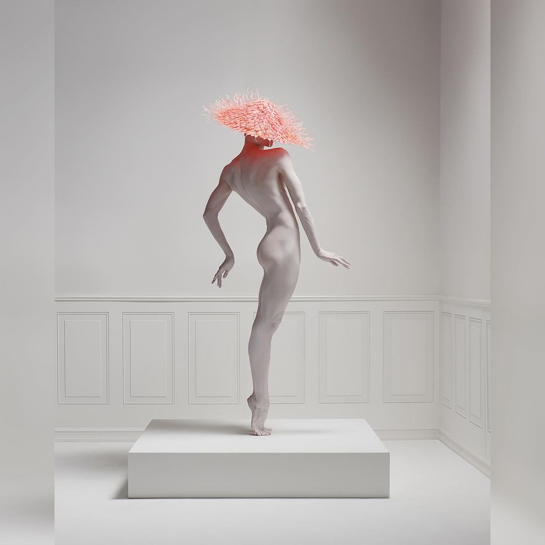 A nude adult woman stands on a white cube in a bright white studio with decorative wall panels, wearing a textured pink headpiece.