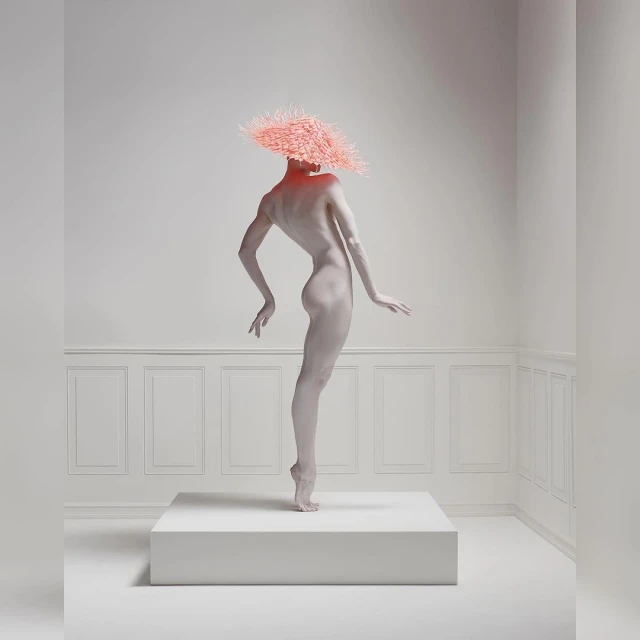A nude adult woman stands on a white cube in a bright white studio with decorative wall panels, wearing a textured pink headpiece.