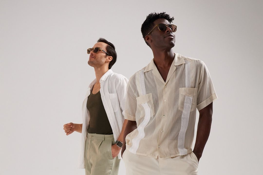 Two men wearing sunglasses and casual shirts pose against a white backdrop in a fashion style shoot with soft lighting and neutral tones.