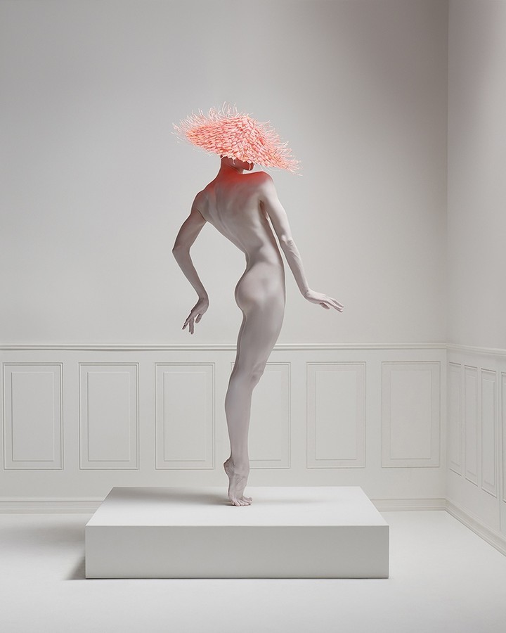 Artistic studio shot of a nude female figure, body painted white, standing on a white cube in an all-white room wearing a pink spiky hat.