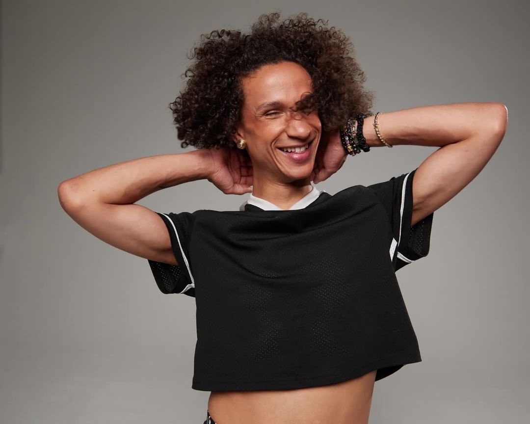 Smiling adult with curly hair poses in front of a grey background, wearing a black mesh crop top and bracelets.