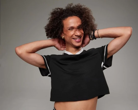 Smiling adult with curly hair poses in front of a grey background, wearing a black mesh crop top and bracelets.