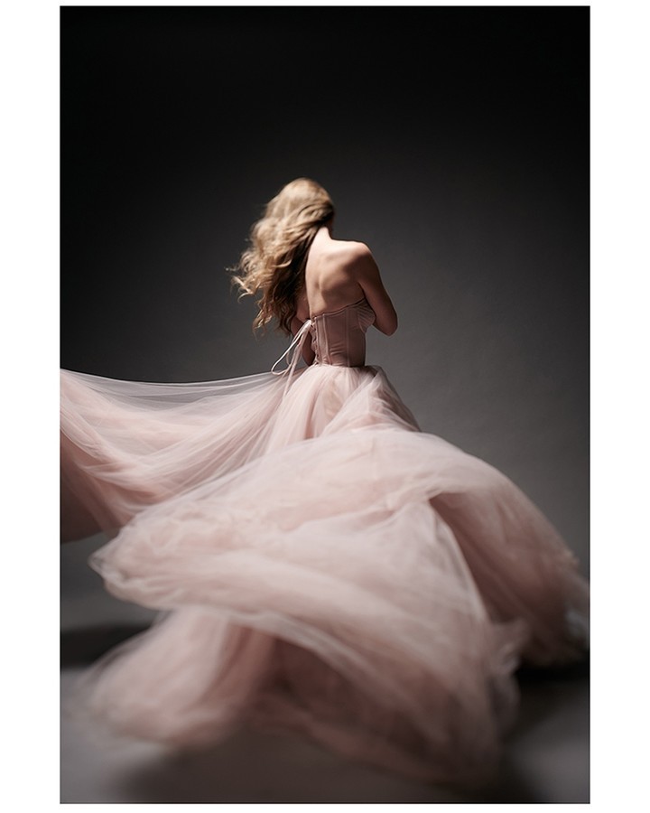 Blonde woman with her back to the camera wearing a pink gown in a photo studio. She is the primary subject of the shot.