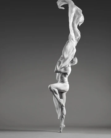Monochromatic dance photography of a lithe male dancer in suspended ballet pose draped with a sheer white fabric, against a grey backdrop.