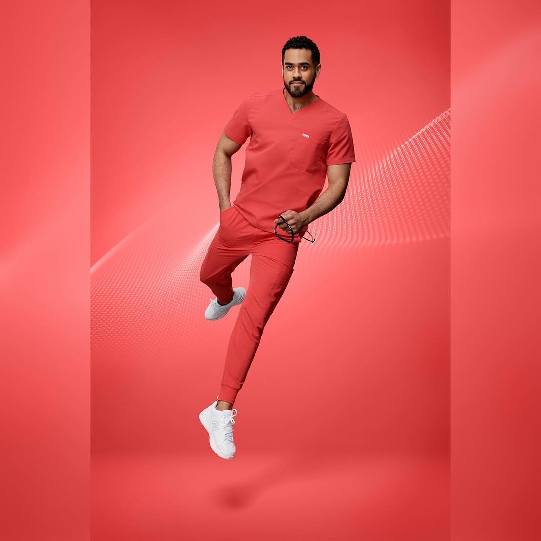 Adult male wearing red scrubs and white sneakers jumps against a red background with graphics. He holds glasses in his left hand.