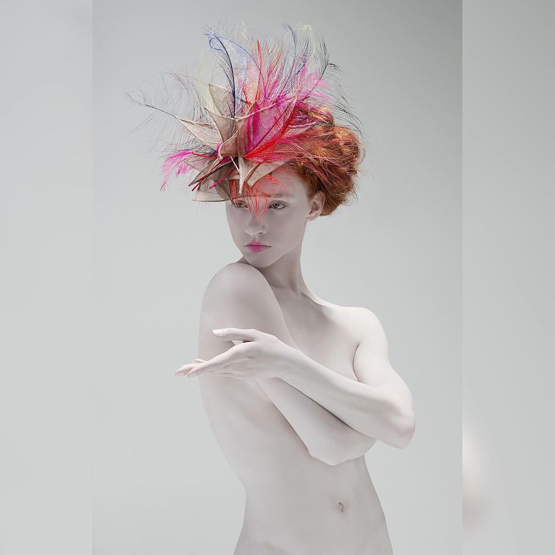 Woman with pale skin and red hair wearing feathered headpiece in front of grey background. Fashion photography. Beauty shot.