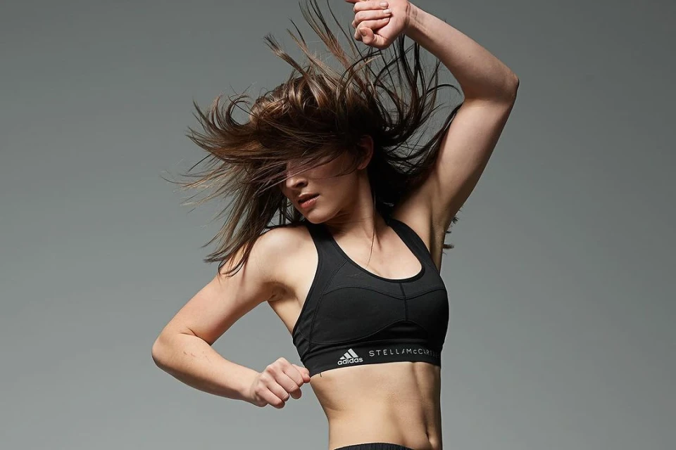 A woman in athletic wear strikes a dynamic pose against a grey backdrop, her hair frozen mid-air. Adidas Stella McCartney branding is visible.