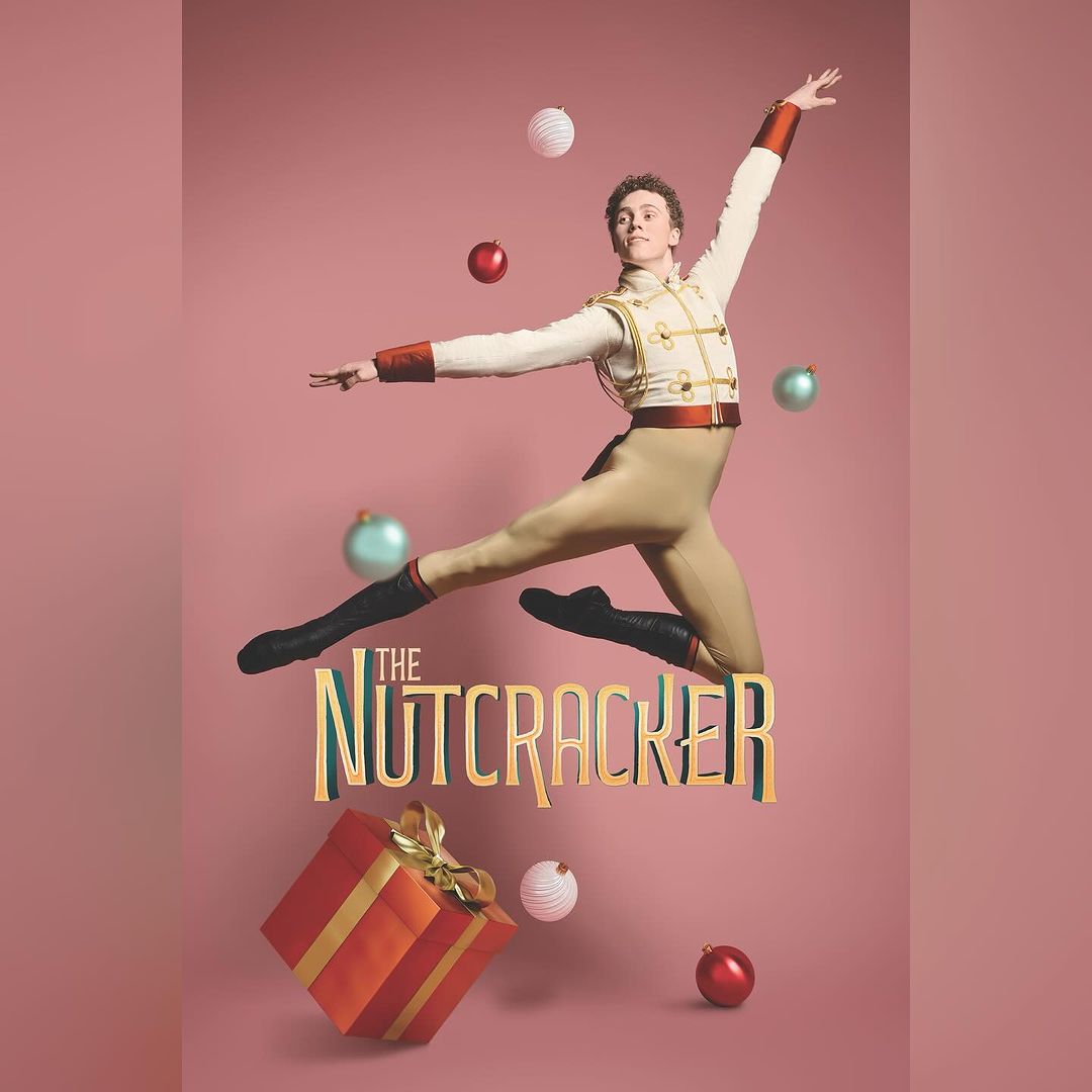 Ballet dancer in the air in a leaping pose, wearing a beige and white Nutcracker prince uniform, on a pink background with Christmas decorations.